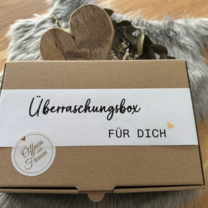 Könnte beinhalten: Eine braune Pappschachtel mit den Worten "Überraschungsbox FÜR DICH" auf einem weißen Etikett. Ein Holzherz und Trockenblumen liegen auf der Schachtel. Ein kleiner Aufkleber mit Text befindet sich auf der Schachtel.