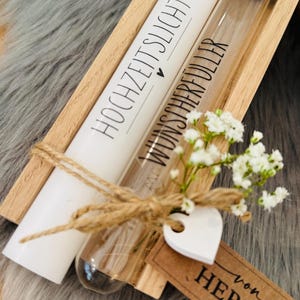 K&ouml;nnte beinhalten: Eine Holzkiste enth&auml;lt eine wei&szlig;e Kerze mit dem Text "HOCHZEITSLICHT" und ein Glasr&ouml;hrchen mit dem Text "WUNSCHERF&Uuml;LLER". Die Artikel sind mit einer Schnurschleife, einem wei&szlig;en Herz und einem Etikett mit "von HERZEN" verziert.