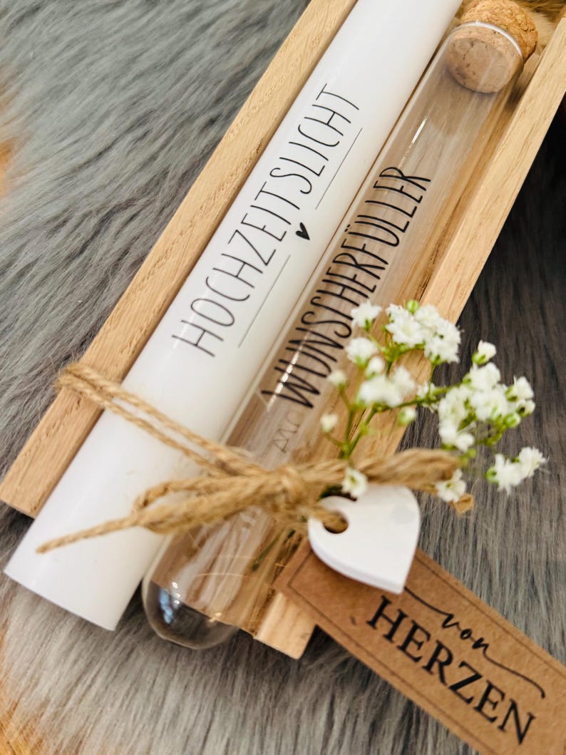 K&ouml;nnte beinhalten: Eine Holzkiste enth&auml;lt eine wei&szlig;e Kerze mit dem Text "HOCHZEITSLICHT" und ein Glasr&ouml;hrchen mit dem Text "WUNSCHERF&Uuml;LLER". Die Artikel sind mit einer Schnurschleife, einem wei&szlig;en Herz und einem Etikett mit "von HERZEN" verziert.