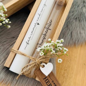 K&ouml;nnte beinhalten: Ein wei&szlig;er zylindrischer Beh&auml;lter und ein Glasfl&auml;schchen mit Korken werden in einem Holztablett pr&auml;sentiert. Der Beh&auml;lter tr&auml;gt die Aufschrift "f&uuml;r immer und ewig" und "Wunscherf&uuml;ller". Ein kleines wei&szlig;es Herz und ein braunes Etikett mit "von HERZEN" sind befestigt.