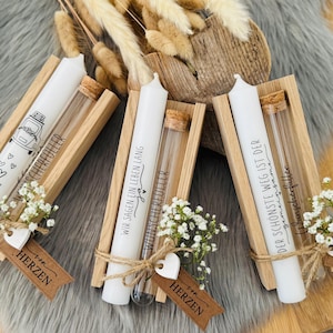 K&ouml;nnte beinhalten: Drei wei&szlig;e Kerzen mit Text und Illustrationen, jeweils in einem Holzhalter mit einem Glasr&ouml;hrchen und kleinen wei&szlig;en Blumen. Die Kerzen sind mit Bindfaden gebunden und haben herzf&ouml;rmige Anh&auml;nger mit der Aufschrift "HERZEN".