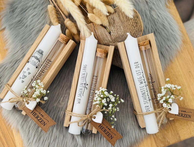 K&ouml;nnte beinhalten: Drei wei&szlig;e Kerzen mit Text und Illustrationen, jeweils in einem Holzhalter mit einem Glasr&ouml;hrchen und kleinen wei&szlig;en Blumen. Die Kerzen sind mit Bindfaden gebunden und haben herzf&ouml;rmige Anh&auml;nger mit der Aufschrift "HERZEN".
