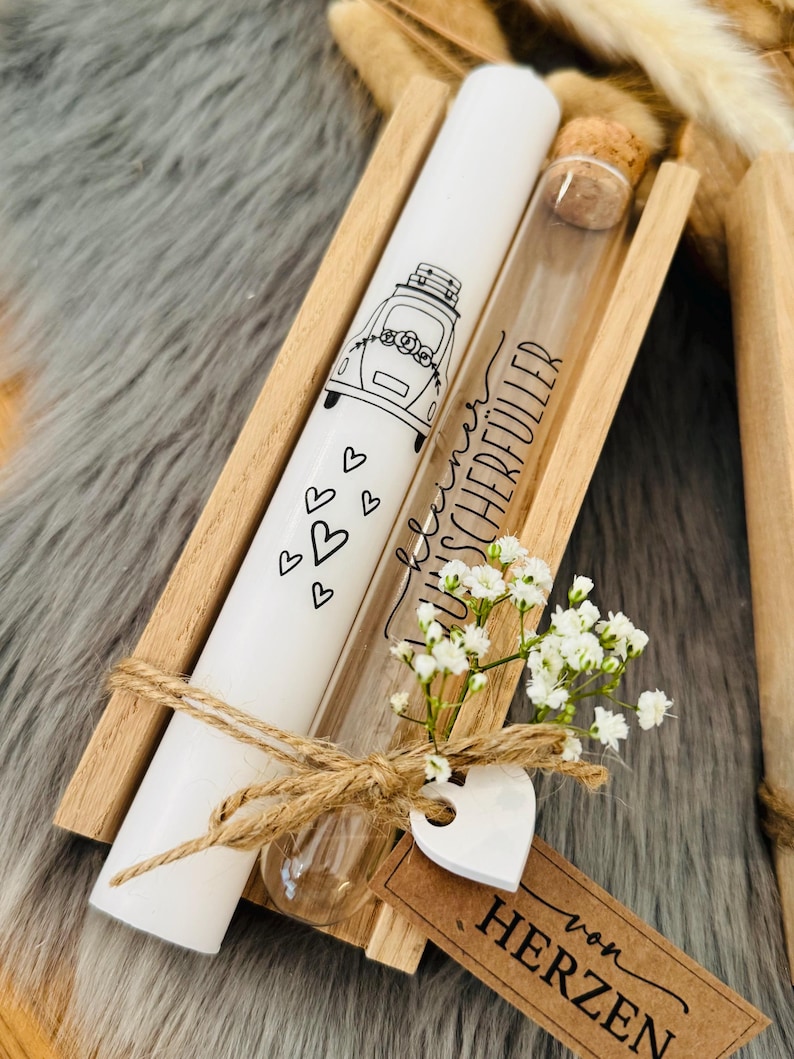 K&ouml;nnte beinhalten: Eine wei&szlig;e Kerze mit einer Autoillustration und Herzen, ein Glasfl&auml;schchen mit Korken und ein kleiner Strau&szlig; wei&szlig;er Blumen werden in einer Holzkiste pr&auml;sentiert. Der Text "von HERZEN" befindet sich auf einem braunen Etikett.