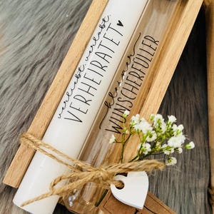 K&ouml;nnte beinhalten: Eine Holzkiste enth&auml;lt eine wei&szlig;e Kerze und ein Glasr&ouml;hrchen, beide mit schwarzer Schrift. Auf der Kerze steht "VERHEIRATET" und auf dem R&ouml;hrchen "WUNSCHERF&Uuml;LLER". Ein kleiner Strau&szlig; wei&szlig;er Blumen und ein herzf&ouml;rmiges Etikett sind mit Bindfaden gebunden.