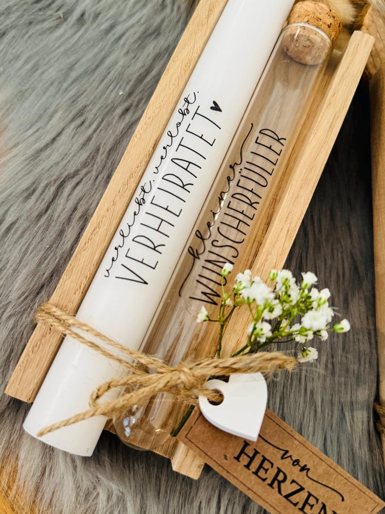 K&ouml;nnte beinhalten: Eine Holzkiste enth&auml;lt eine wei&szlig;e Kerze und ein Glasr&ouml;hrchen, beide mit schwarzer Schrift. Auf der Kerze steht "VERHEIRATET" und auf dem R&ouml;hrchen "WUNSCHERF&Uuml;LLER". Ein kleiner Strau&szlig; wei&szlig;er Blumen und ein herzf&ouml;rmiges Etikett sind mit Bindfaden gebunden.