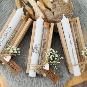K&ouml;nnte beinhalten: Drei wei&szlig;e Kerzen mit Text in Holzhaltern, jeweils mit einem Glasr&ouml;hrchen und einem Korken. Die Kerzen sind mit Bindf&auml;den, kleinen wei&szlig;en Blumen und herzf&ouml;rmigen Anh&auml;ngern verziert. Der Text auf den Kerzen enth&auml;lt S&auml;tze wie "Verheiratet".