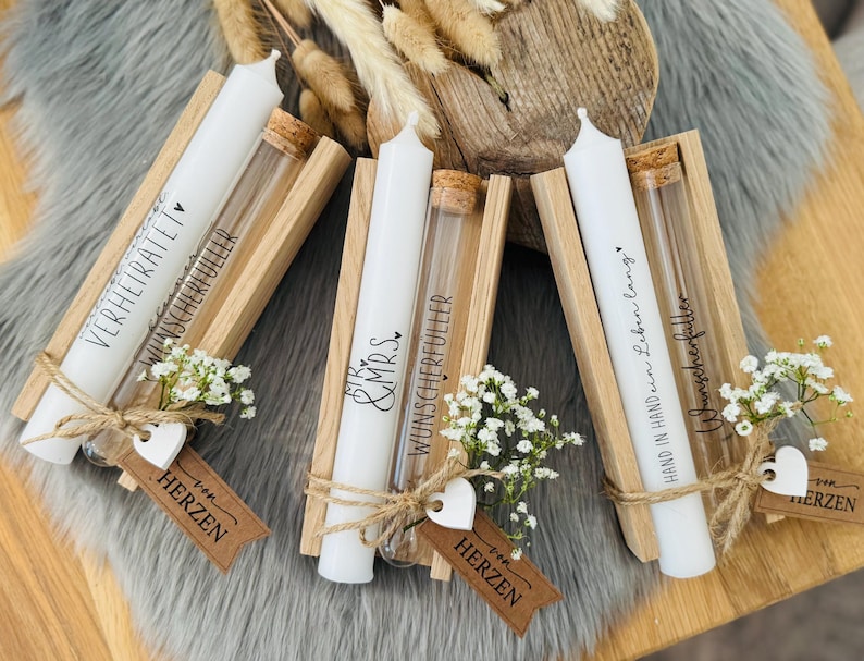 K&ouml;nnte beinhalten: Drei wei&szlig;e Kerzen mit Text in Holzhaltern, jeweils mit einem Glasr&ouml;hrchen und einem Korken. Die Kerzen sind mit Bindf&auml;den, kleinen wei&szlig;en Blumen und herzf&ouml;rmigen Anh&auml;ngern verziert. Der Text auf den Kerzen enth&auml;lt S&auml;tze wie "Verheiratet".