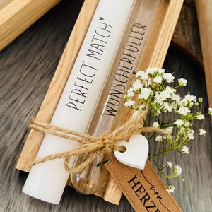 K&ouml;nnte beinhalten: Eine Holzkiste enth&auml;lt eine wei&szlig;e Kerze und ein Glasr&ouml;hrchen mit Korken. Auf der Kerze steht "PERFECT MATCH" und auf dem R&ouml;hrchen "WUNSCHERF&Uuml;LLER". Ein wei&szlig;es Herz-Etikett und ein braunes Etikett mit Text sind mit Bindfaden gebunden.