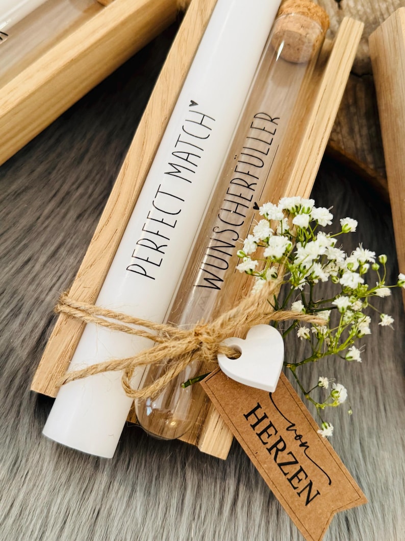 K&ouml;nnte beinhalten: Eine Holzkiste enth&auml;lt eine wei&szlig;e Kerze und ein Glasr&ouml;hrchen mit Korken. Auf der Kerze steht "PERFECT MATCH" und auf dem R&ouml;hrchen "WUNSCHERF&Uuml;LLER". Ein wei&szlig;es Herz-Etikett und ein braunes Etikett mit Text sind mit Bindfaden gebunden.