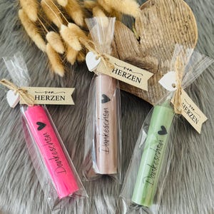 Könnte beinhalten: Drei verpackte Kerzen in Rosa, Beige und Grün, jede mit einem Herz und dem Wort "Dankeschen" bedruckt. Die Kerzen sind mit Bindfaden und einem Etikett mit der Aufschrift "von Herzen" gebunden. Der Hintergrund umfasst Trockenblumen und ein Holzherz.