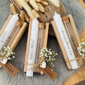 K&ouml;nnte beinhalten: Drei wei&szlig;e Kerzen mit Text in Holzhaltern, jeweils mit einem kleinen Glasr&ouml;hrchen und einem Zweig wei&szlig;er Blumen. Die Kerzen sind mit Bindfaden gebunden und haben kleine Anh&auml;nger.