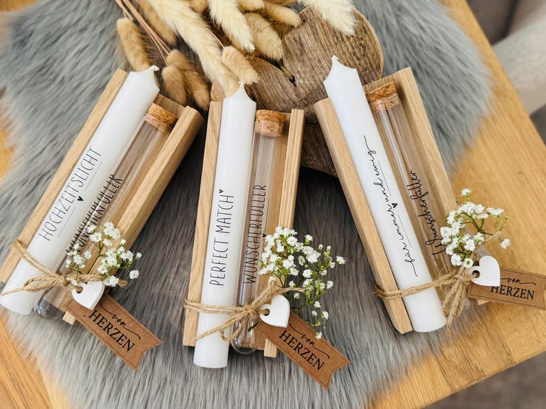 K&ouml;nnte beinhalten: Drei wei&szlig;e Kerzen mit Text in Holzhaltern, jeweils mit einem kleinen Glasr&ouml;hrchen und einem Zweig wei&szlig;er Blumen. Die Kerzen sind mit Bindfaden gebunden und haben kleine Anh&auml;nger.