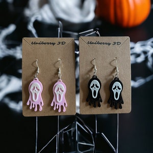 Peut inclure: Deux paires de boucles d'oreilles sur une carte marron avec le texte "uniberry 3D". Les boucles d'oreilles sont en forme de visage criant avec de la peinture qui coule. Une paire est rose et l'autre est noire.