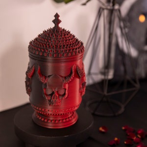 Peut inclure: Un pot en céramique rouge avec un motif de crâne et un couvercle à pointes. Le pot est décoré de détails complexes et a une esthétique gothique sombre.