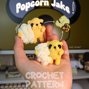 May include: Two small yellow crocheted figures with black eyes, each holding a cluster of white popcorn-like shapes. The text Popcorn Jake and CROCHET PATTERN is visible, along with a small illustration of the character and popcorn.
