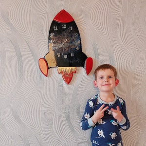 Op de afbeelding: Een jonge jongen in een blauwe pyjama met een wit patroon staat voor een witte muur met een raketvormige klok. De klok heeft een zwarte wijzerplaat met witte cijfers en een rood en geel raketontwerp.