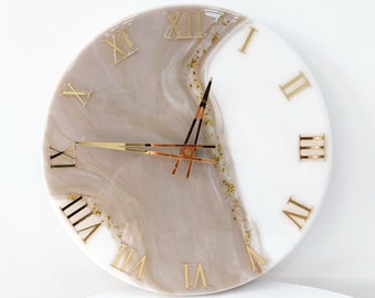 May include: A white and beige resin clock with gold Roman numeral markings. The clock face has a swirling pattern and gold glitter accents.
