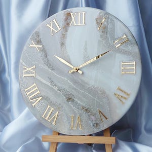 Puede incluir: Reloj de pared redondo con números romanos y manecillas doradas. La esfera del reloj tiene un diseño veteado blanco y gris con detalles dorados. El reloj se exhibe sobre un pequeño caballete de madera.