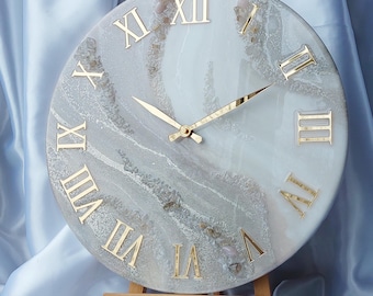 May include: Round wall clock with gold Roman numerals and hands. The clock face has a white and gray marbled design with gold accents. The clock is displayed on a small wooden easel.
