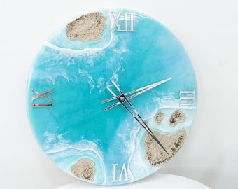 Puede incluir: Un reloj redondo con una cara de resina azul y blanca que representa una escena de playa con olas y arena. La esfera del reloj tiene marcas de números romanos y manecillas plateadas.