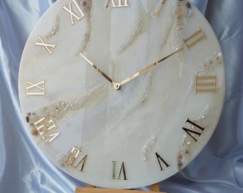 May include: A round wall clock with a white and beige marbled design. The clock features gold Roman numerals and hands. The clock is displayed on a small wooden easel. The clock's diameter is approximately 12 inches.