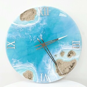 May include: A blue and white resin clock with Roman numeral markings and a beach scene design. The clock face features a blue ocean with white waves and brown sand islands.