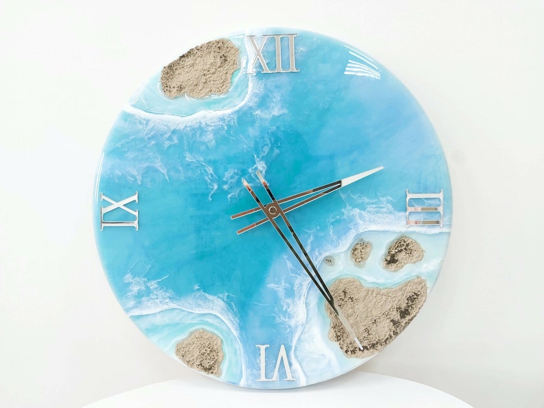 May include: A blue and white resin clock with Roman numeral markings and a beach scene design. The clock face features a blue ocean with white waves and brown sand islands.
