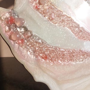 May include: Close-up of a decorative resin art piece with a flowing, organic shape. The piece features layers of white and pink resin, embedded with clear and pink stones, and gold flecks.