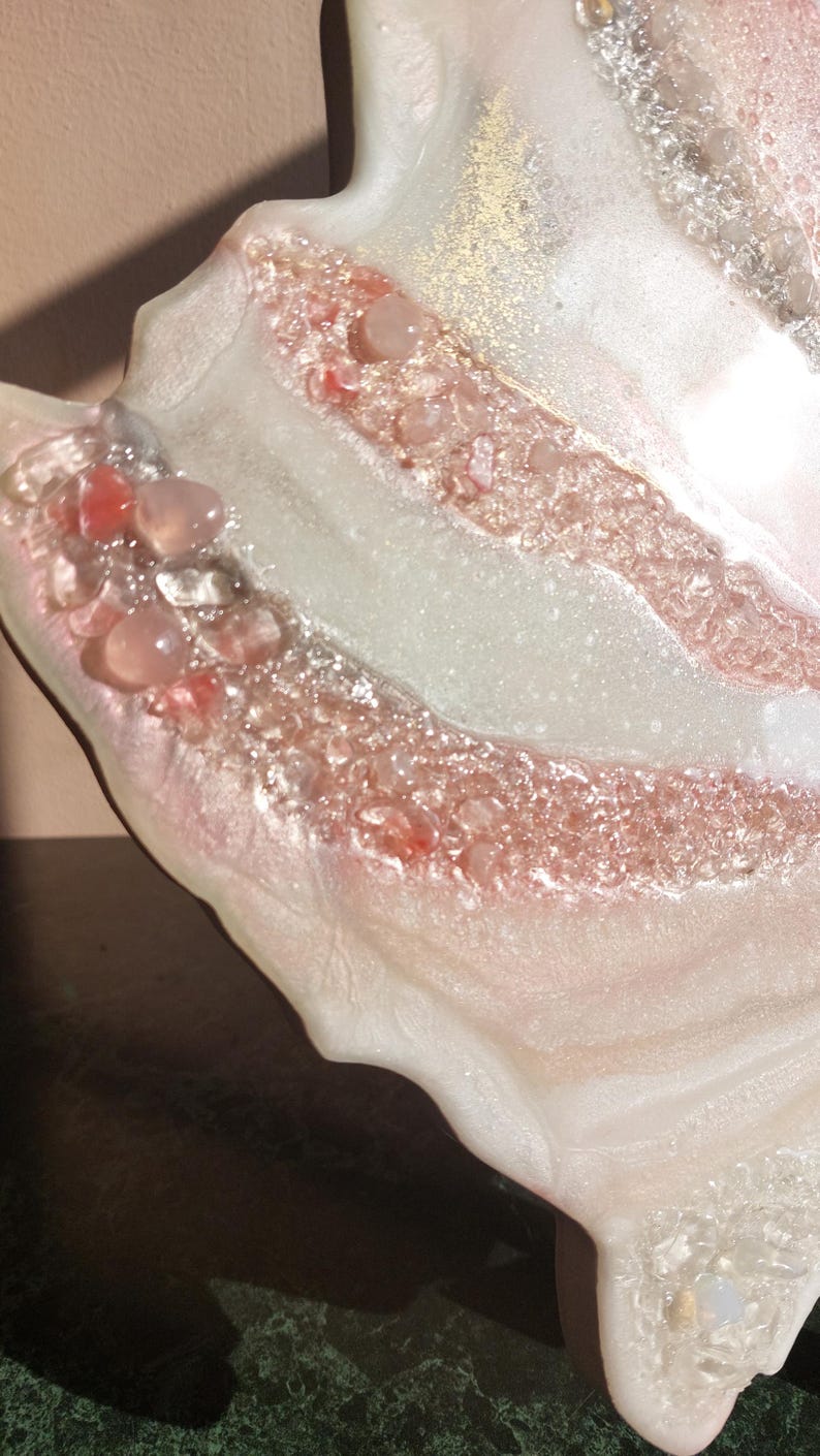 May include: Close-up of a decorative resin art piece with a flowing, organic shape. The piece features layers of white and pink resin, embedded with clear and pink stones, and gold flecks.