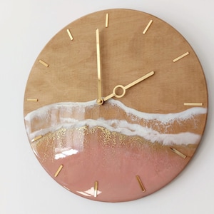 May include: A round wooden clock with gold hands and a pink, white and gold resin design that resembles a beach scene.