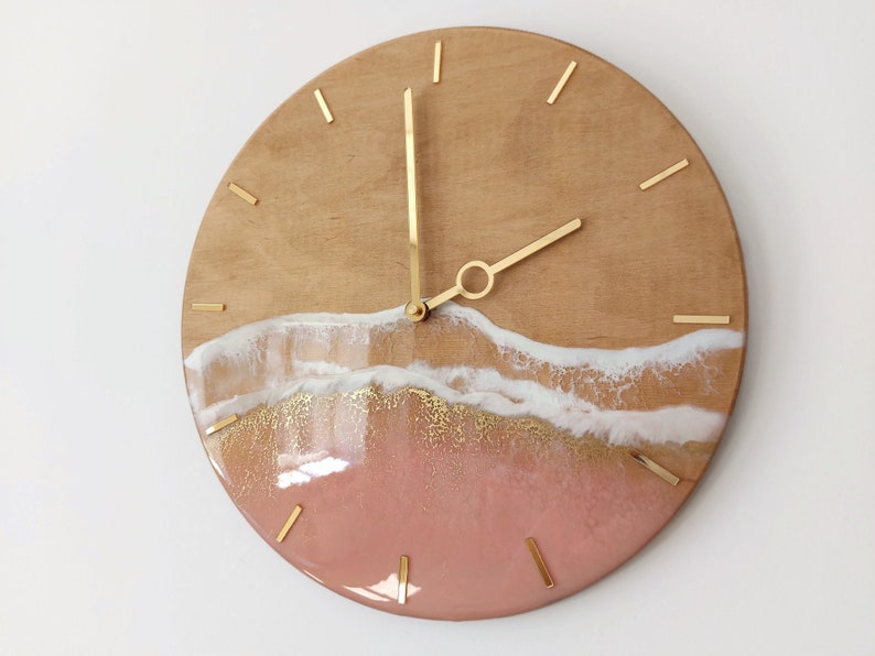 May include: A round wooden clock with gold hands and a pink, white, and gold resin design that resembles a beach scene.