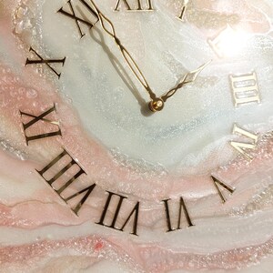 May include: A decorative wall clock with a unique design. The clock face features Roman numerals in gold against a white, pink, and gray marbled background. The clock hands are gold. The clock is displayed on a wooden easel.