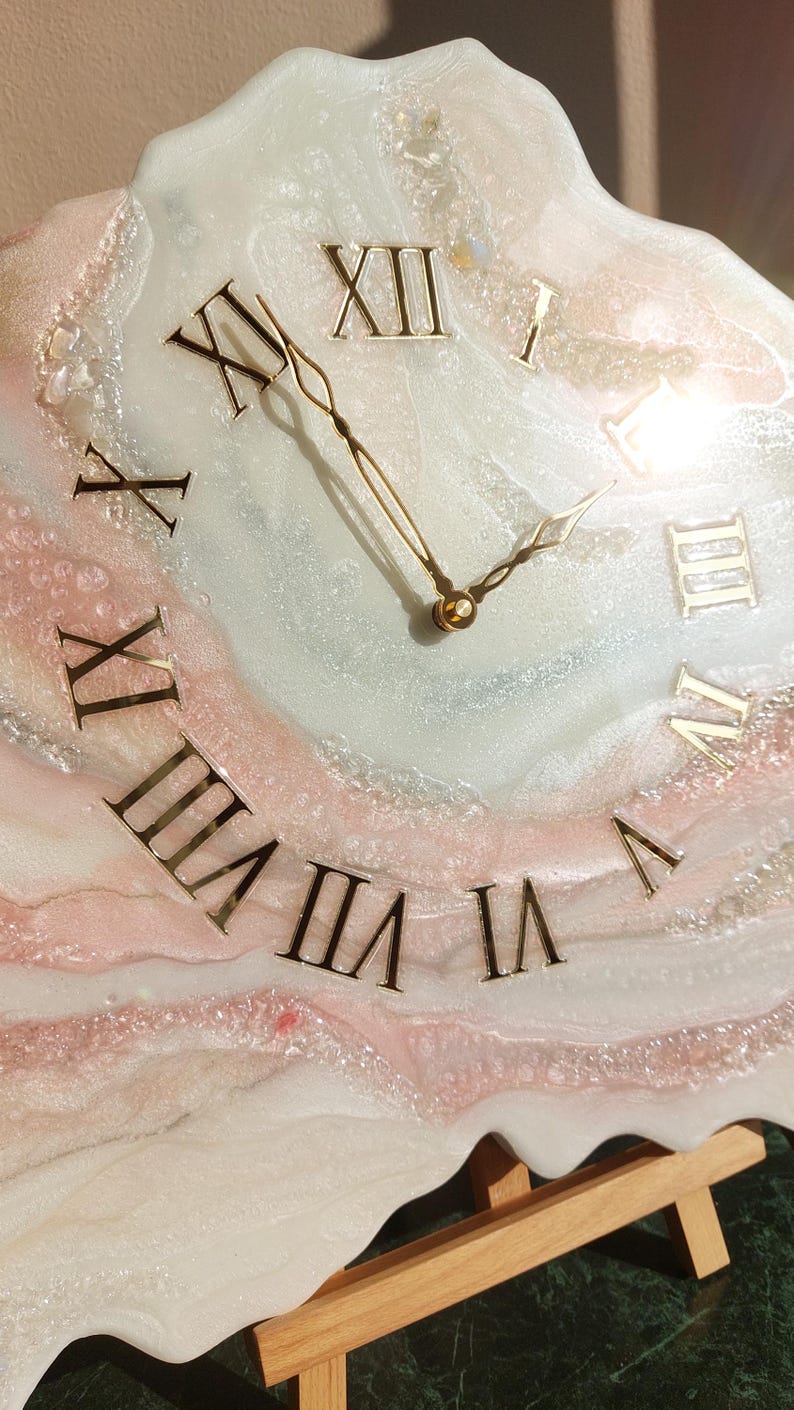 May include: A decorative wall clock with a unique design. The clock face features Roman numerals in gold against a white, pink, and gray marbled background. The clock hands are gold. The clock is displayed on a wooden easel.