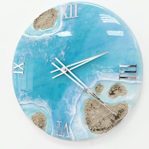 May include: A round blue and white resin clock with Roman numeral markings. The clock face features a design resembling a beach scene with sand and waves.