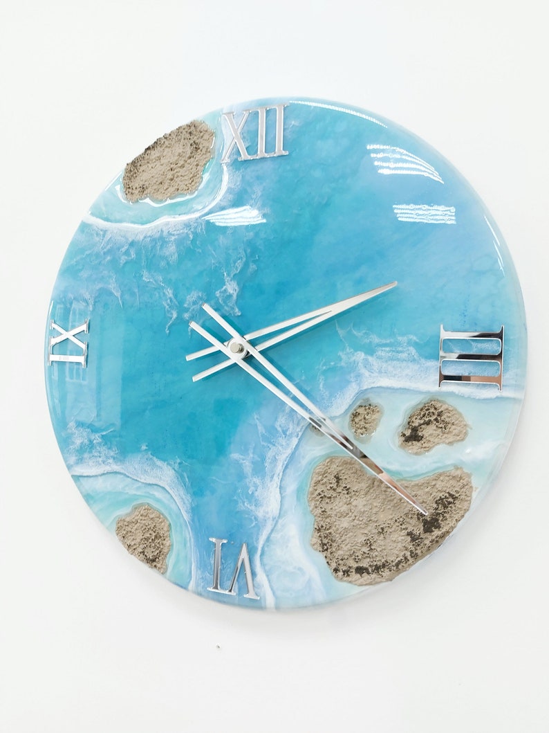 May include: A round blue and white resin clock with Roman numeral markings. The clock face features a design resembling a beach scene with sand and waves.