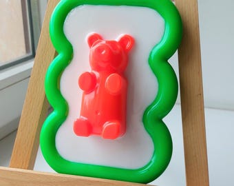 Puede incluir: Un juguete rojo con forma de oso de goma sobre un fondo blanco, enmarcado por un borde verde ondulado. El juguete se exhibe en un pequeño caballete de madera, adecuado para el juego de los niños o la decoración de la habitación.