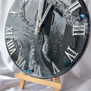 May include: A round, black and silver wall clock with Roman numerals. The clock face features a swirling, abstract design with embedded crystals and a glossy finish. The clock is displayed on a small wooden easel.