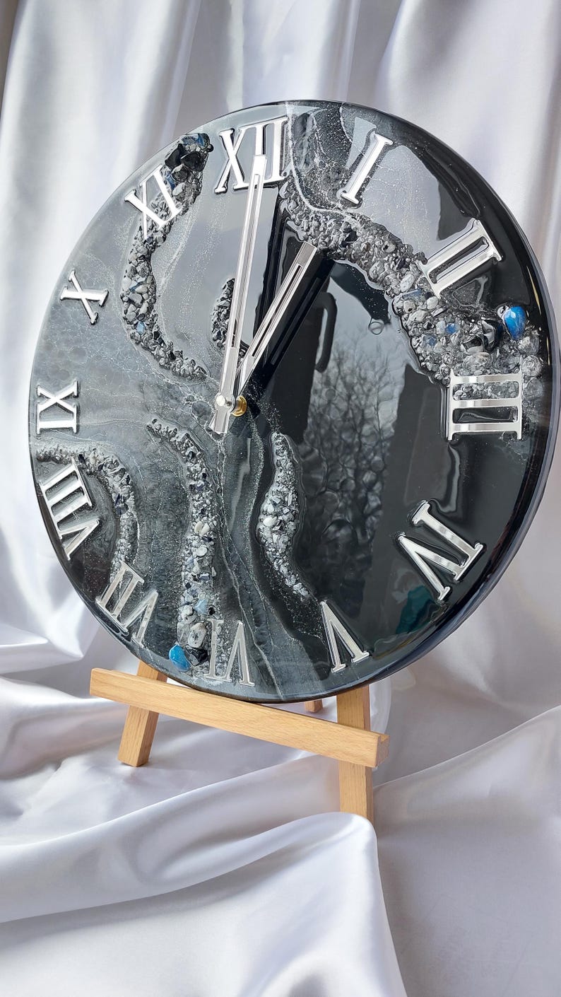 May include: A round, black and silver wall clock with Roman numerals. The clock face features a swirling, abstract design with embedded crystals and a glossy finish. The clock is displayed on a small wooden easel.
