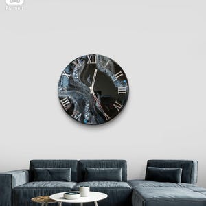 May include: A round wall clock with a black face and Roman numerals. The clock's face features a swirling, abstract design in shades of gray, white, and blue. The clock hands are silver. The clock is mounted on a white wall above a blue sectional sofa.