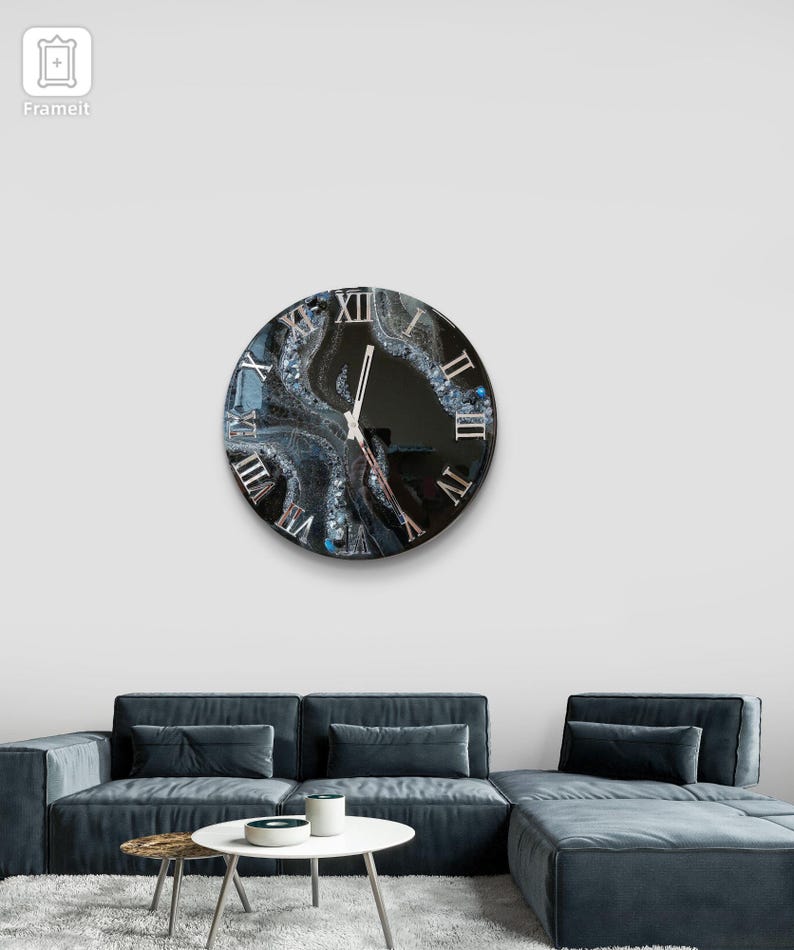 May include: A round wall clock with a black face and Roman numerals. The clock's face features a swirling, abstract design in shades of gray, white, and blue. The clock hands are silver. The clock is mounted on a white wall above a blue sectional sofa.