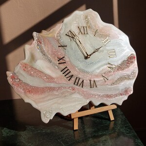 May include: A unique, handcrafted clock with a resin finish. The clock face features Roman numerals and gold-colored hands. The clock is in shades of white, pink, and silver, and is displayed on a small wooden easel.