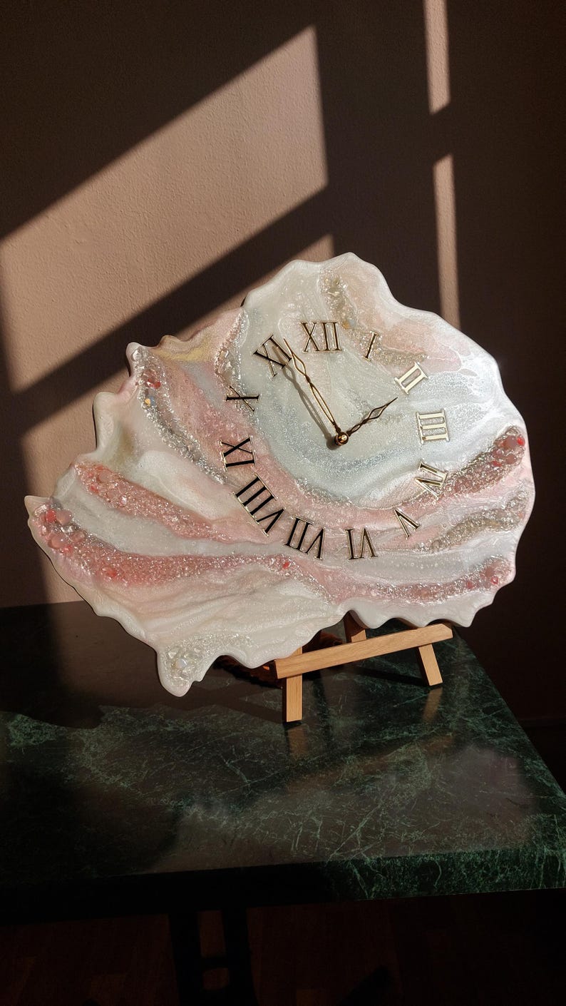 May include: A unique, handcrafted clock with a resin finish. The clock face features Roman numerals and gold-colored hands. The clock is in shades of white, pink, and silver, and is displayed on a small wooden easel.