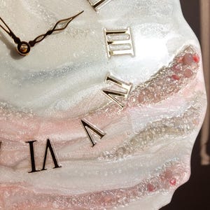 May include: A decorative wall clock with a unique, abstract design. The clock face features a swirling pattern of white, pink, and silver resin, with gold Roman numerals. The clock hands are gold. The clock is displayed on a wooden easel.