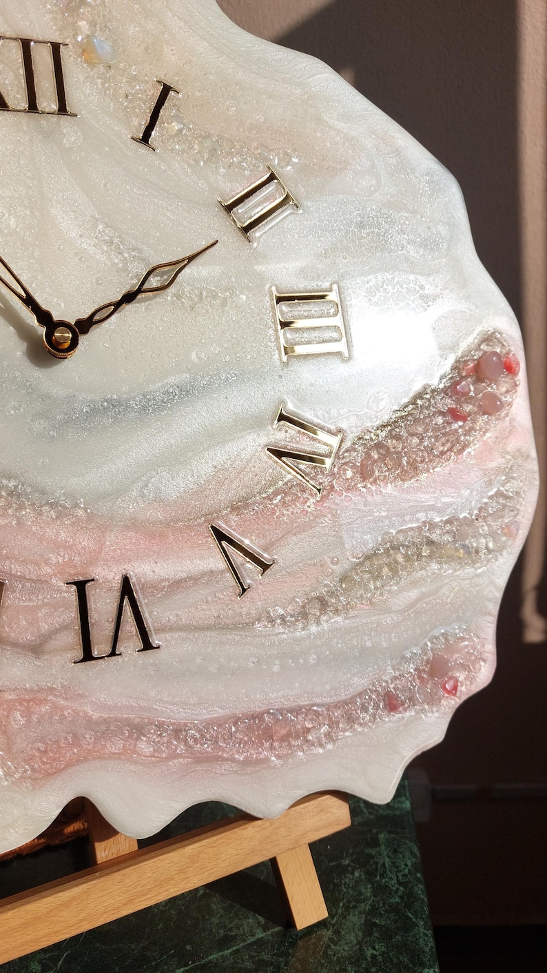 May include: A decorative wall clock with a unique, abstract design. The clock face features a swirling pattern of white, pink, and silver resin, with gold Roman numerals. The clock hands are gold. The clock is displayed on a wooden easel.