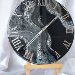 May include: A round, black and silver resin clock with Roman numerals. The clock face features a swirling design with silver and gray accents, and is supported by a small wooden easel. The clock hands are silver.