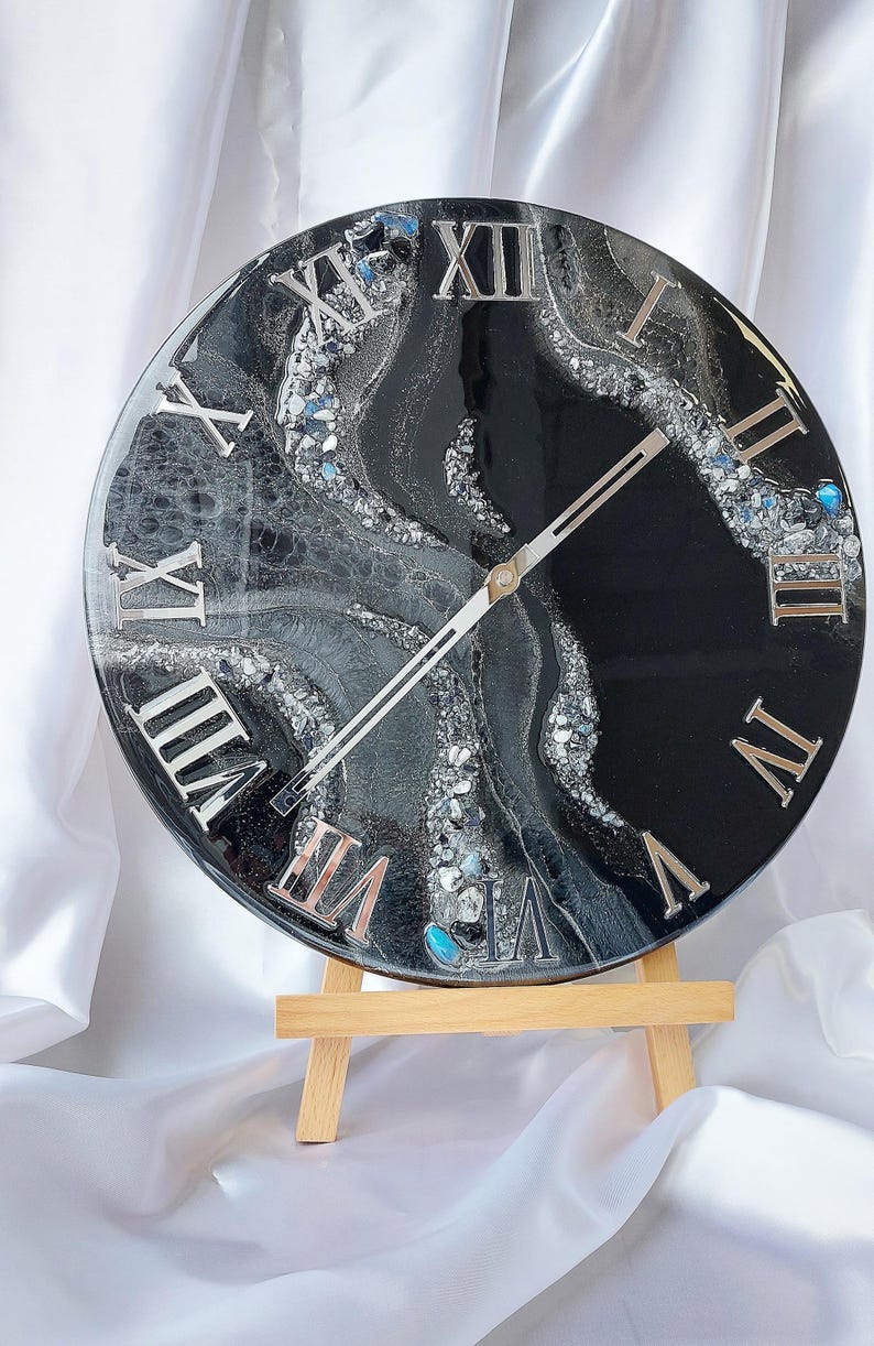 May include: A round, black and silver resin clock with Roman numerals. The clock face features a swirling design with silver and gray accents, and is supported by a small wooden easel. The clock hands are silver.