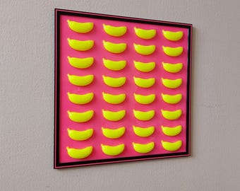 May include: A framed artwork featuring a vibrant pink background with a grid of bright yellow, banana-shaped forms. The artwork is encased in a black frame with a thin pink border, mounted on a neutral-toned wall.