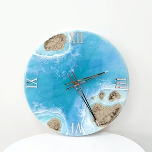 May include: A round blue and white resin clock with Roman numeral markings. The clock face features a design of blue water with white waves and brown sand.