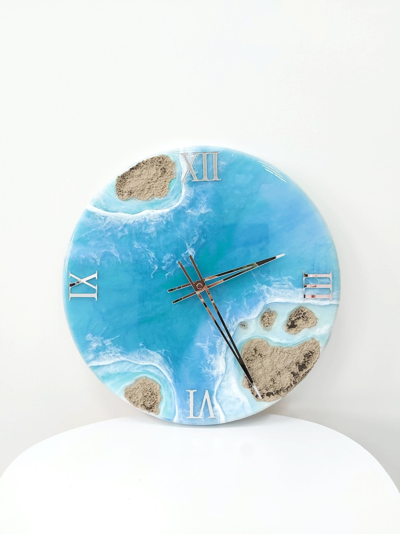 May include: A round blue and white resin clock with Roman numeral markings. The clock face features a design of blue water with white waves and brown sand.