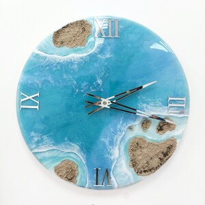 May include: A round blue and white resin clock with Roman numeral markings and silver hands. The clock face features a design of ocean waves and sand.